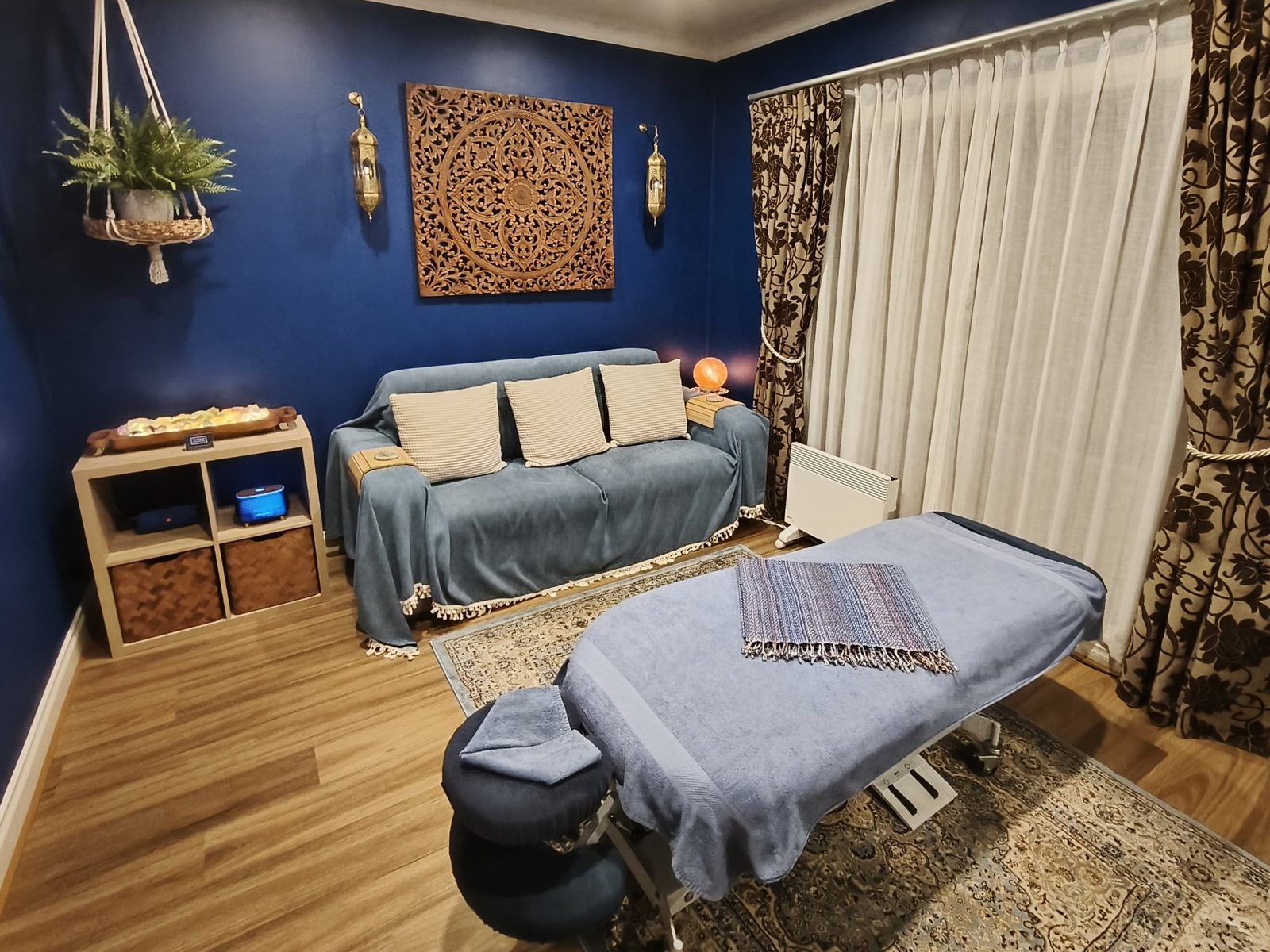The Tempo Bodywork treatment room: deep blue walls, an electric treatment table on a Persian rug, sheer curtains and soft lamplight